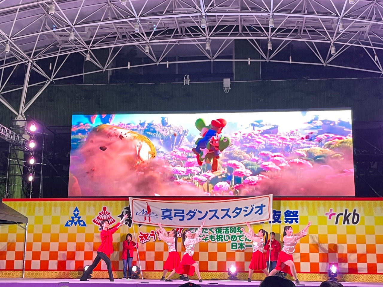 前夜祭inお祭り本舞台　横断幕の前でポーズをとっています。バックのマリオの広告がかわいい