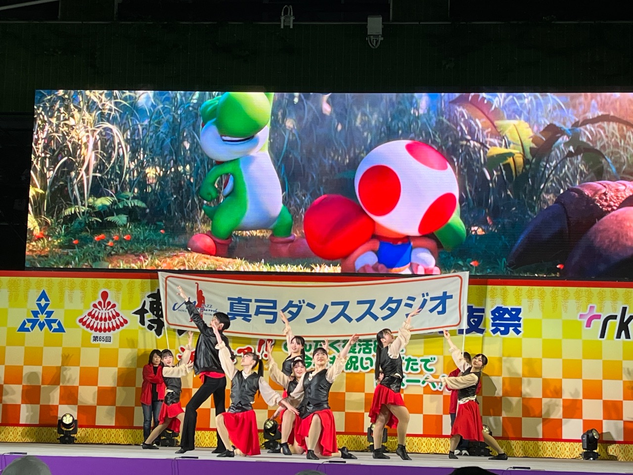 前夜祭inお祭り本舞台　横断幕の前でポーズをとっています。バックのマリオの広告がかわいい