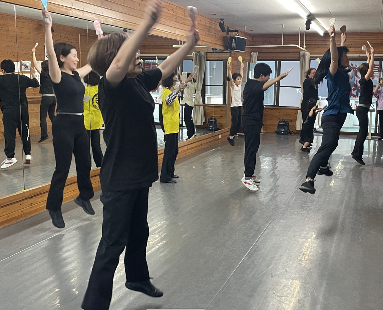 練習スタジオで8人ほどが曲に合わせて両手足を広げて飛び上がっている